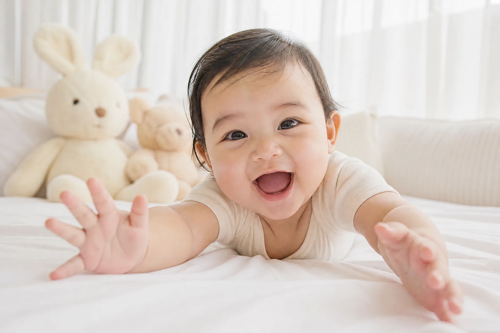 Laughing baby lying on a white bed reaching both open hands toward the camera, soft plush toys and sheer curtains behind