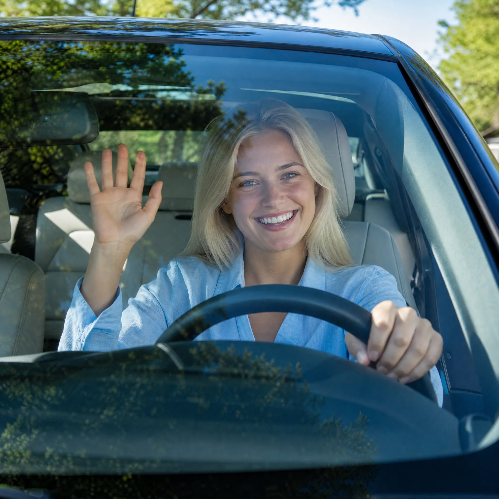 Young blonde woman laughing and waving from the driver’s seat, seen through the windshield on a sunny day
