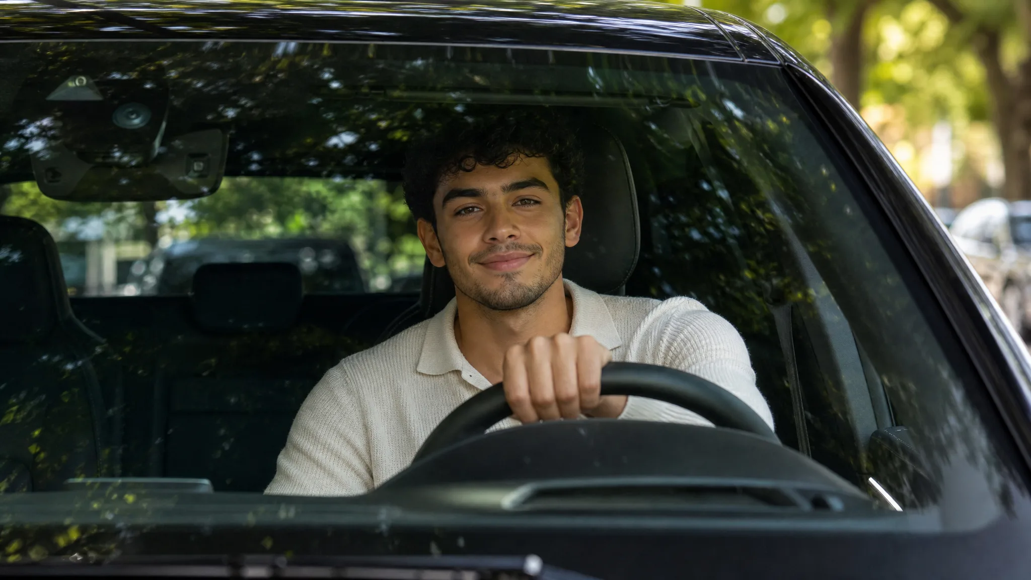 Young man with dark curly hair smiling at the wheel, seen through the windshield of a black car under street trees