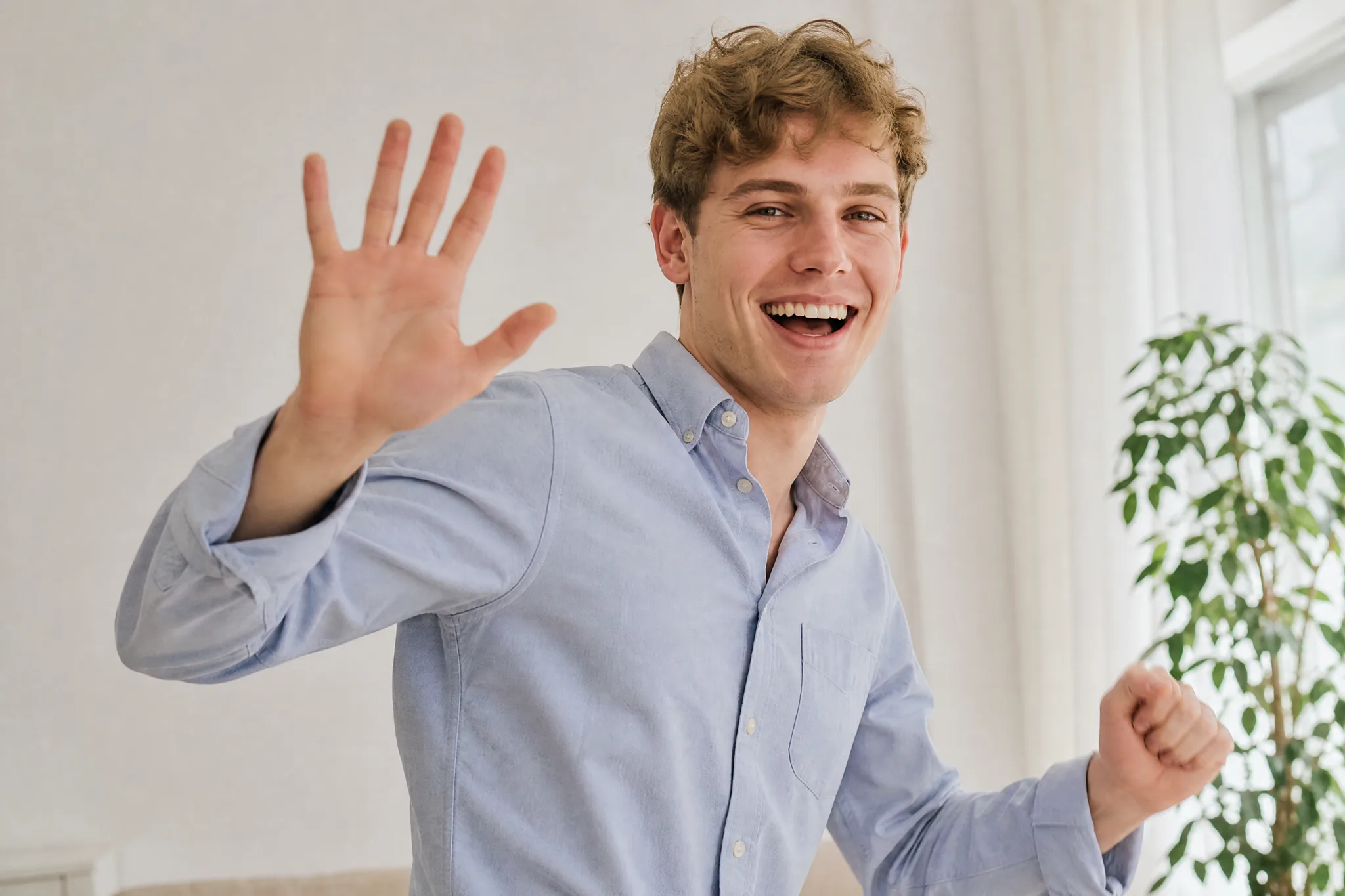 AI Face Dance Generator: A Dance in Time with the Music Young man with wavy brown hair waving at the camera and laughing in a bright white room beside a window