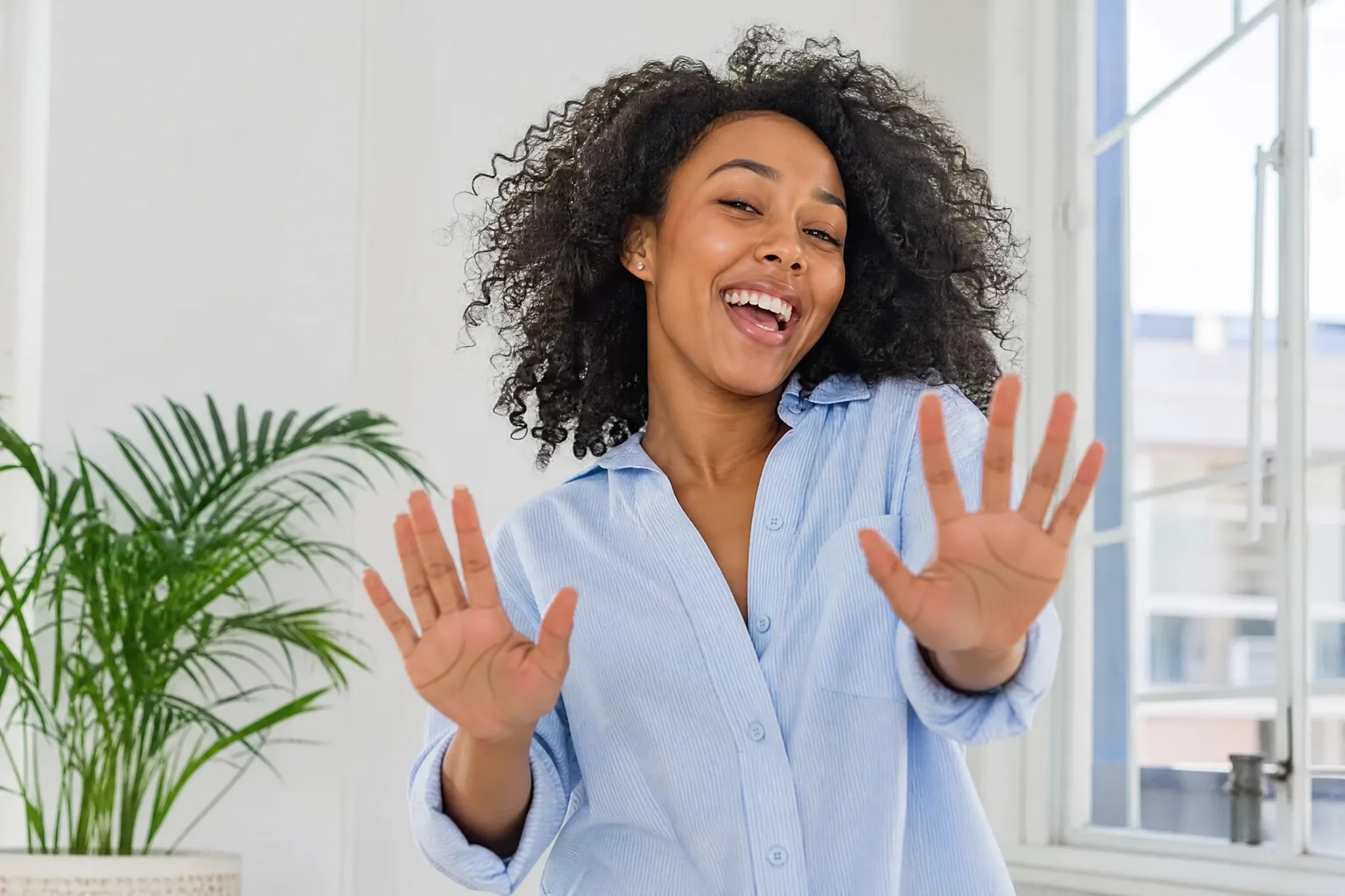 AI Face Dance Generator: Hand-Gesture Moves Young woman with curly black hair grinning and holding both open palms up toward the camera in a bright white room