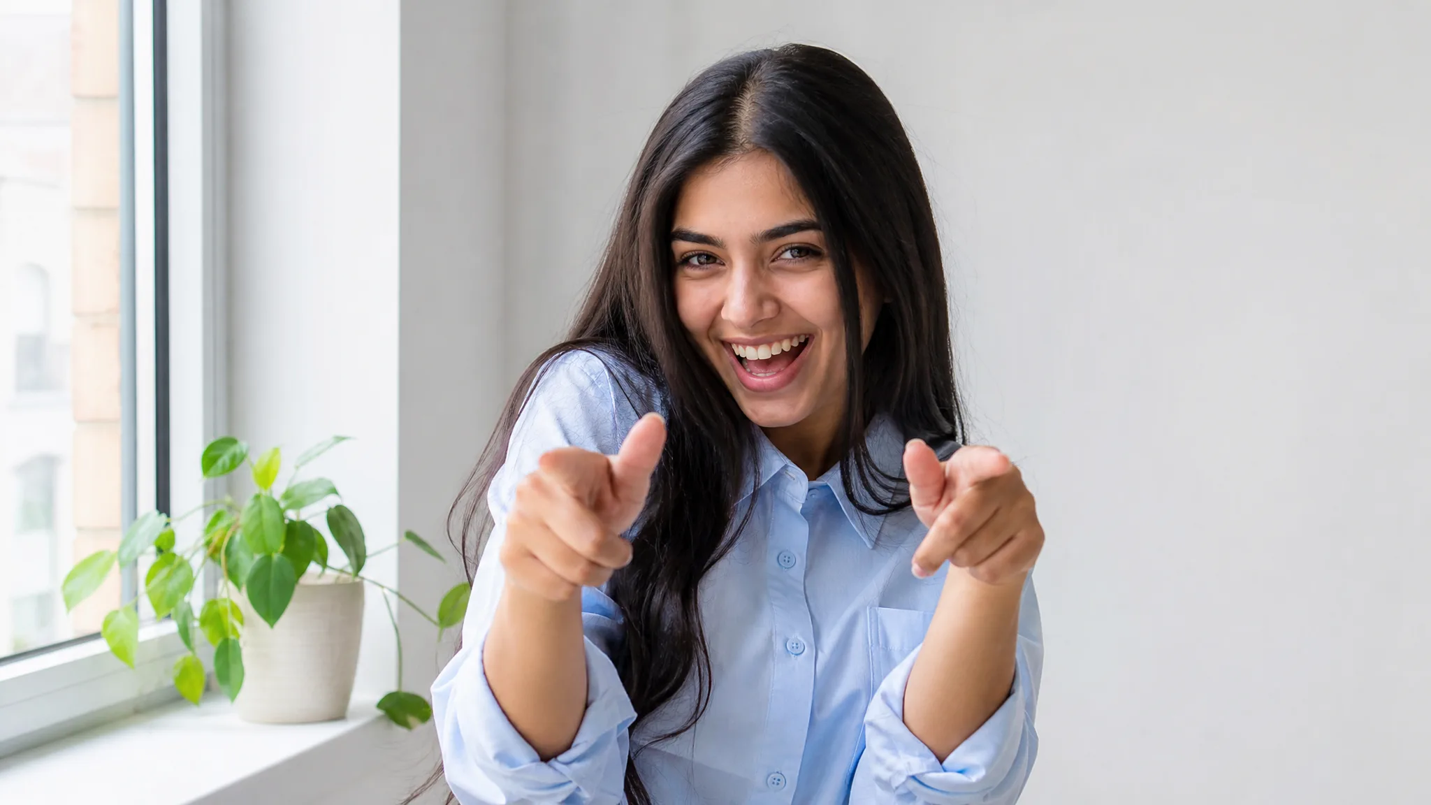 Young woman with long dark hair pointing both index fingers at the camera with a playful grin in a bright white room