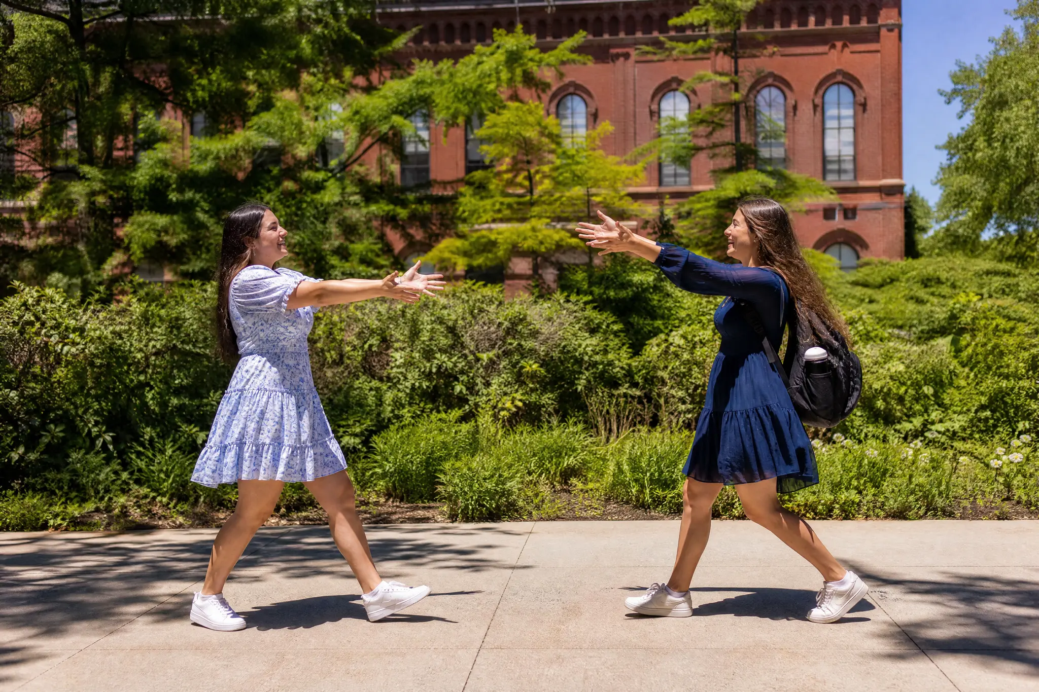 Hugging Video AI: Left and Right in Their Places Two young women walking toward each other with open arms on a sunny walkway in front of a red-brick building