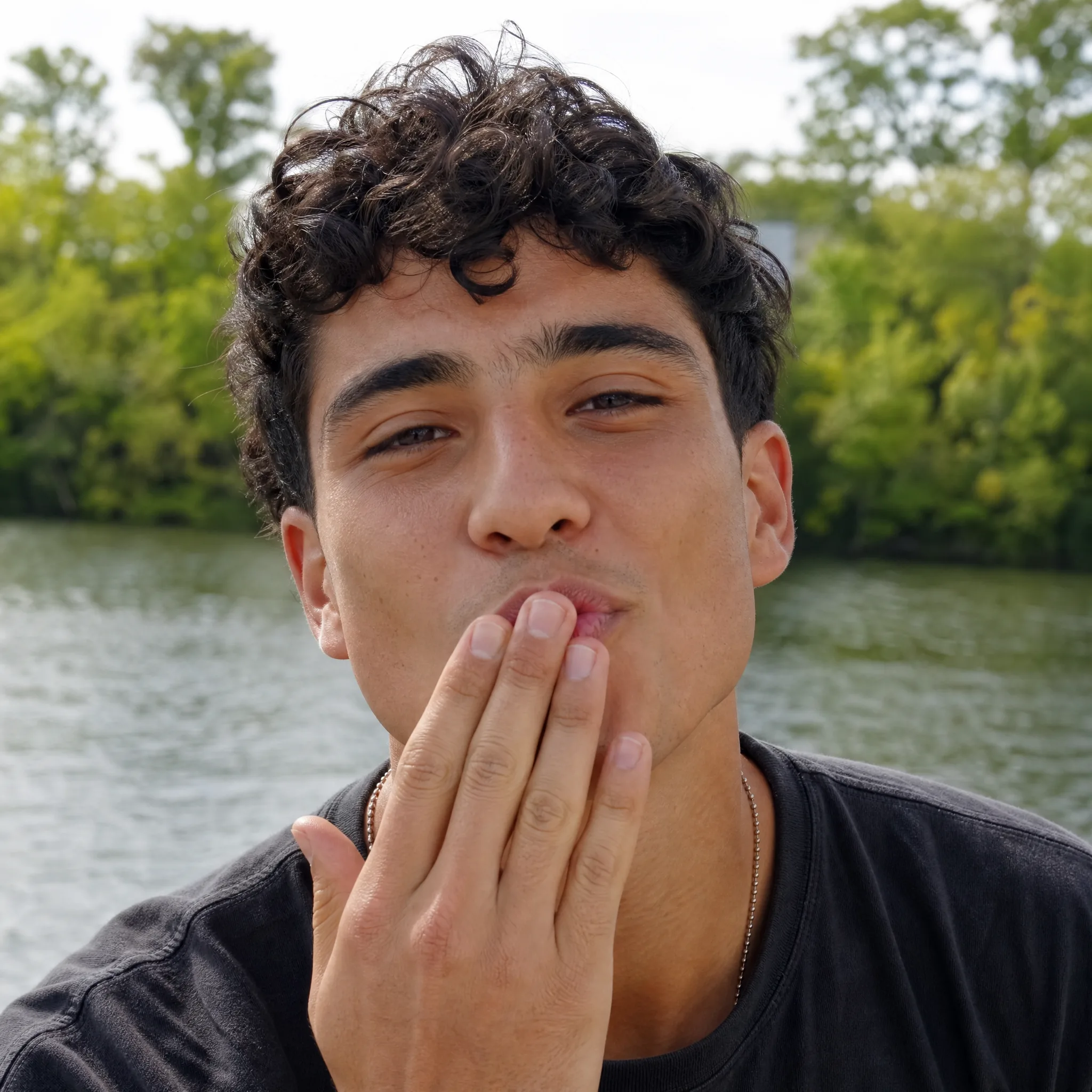 A Romantic Gesture for Your Message Young man with curly hair with his fingertips at his lips, about to blow a kiss on a sunny riverside