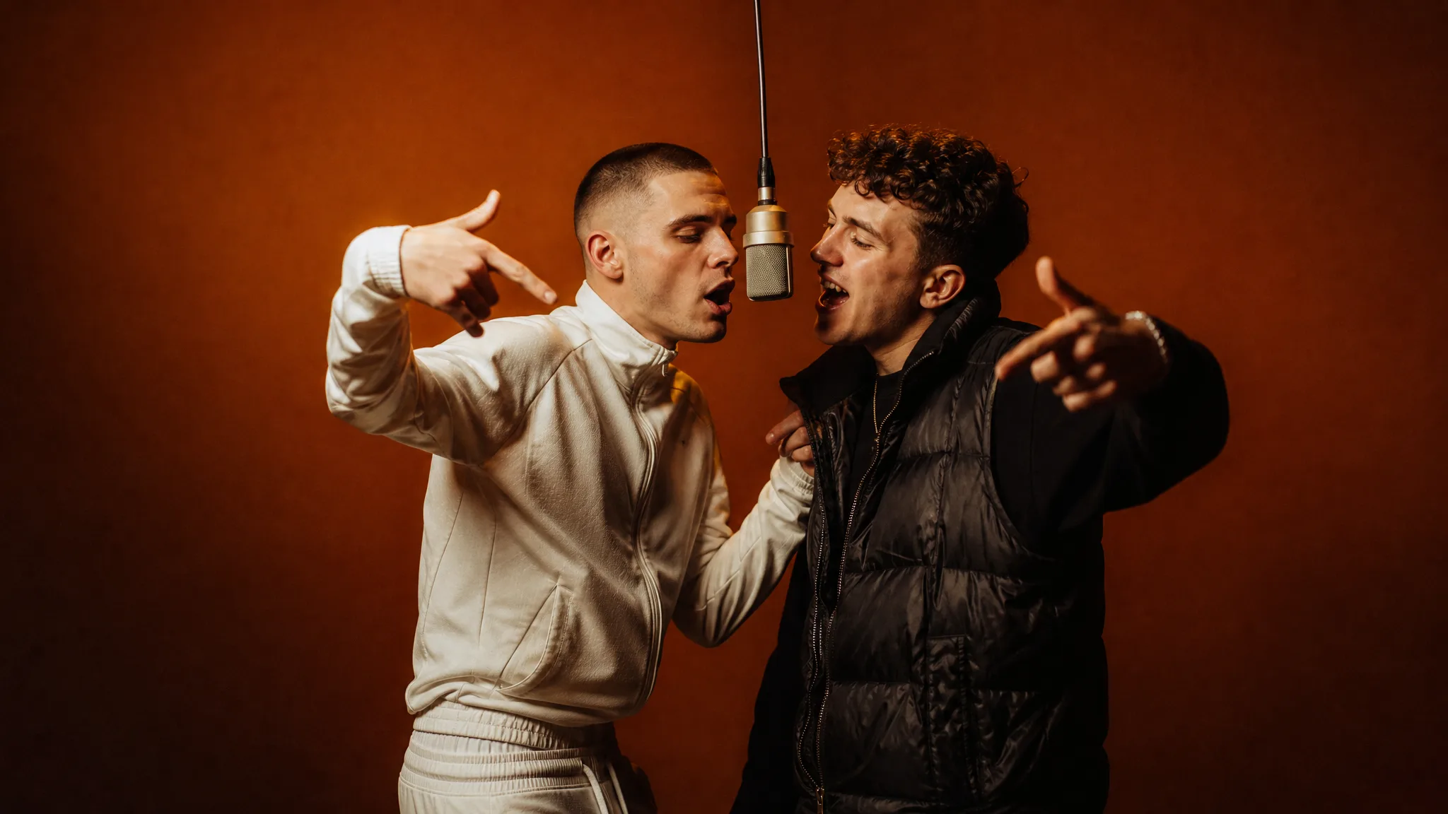Two young men in a white tracksuit and a black puffer vest rapping into a hanging microphone in an orange studio