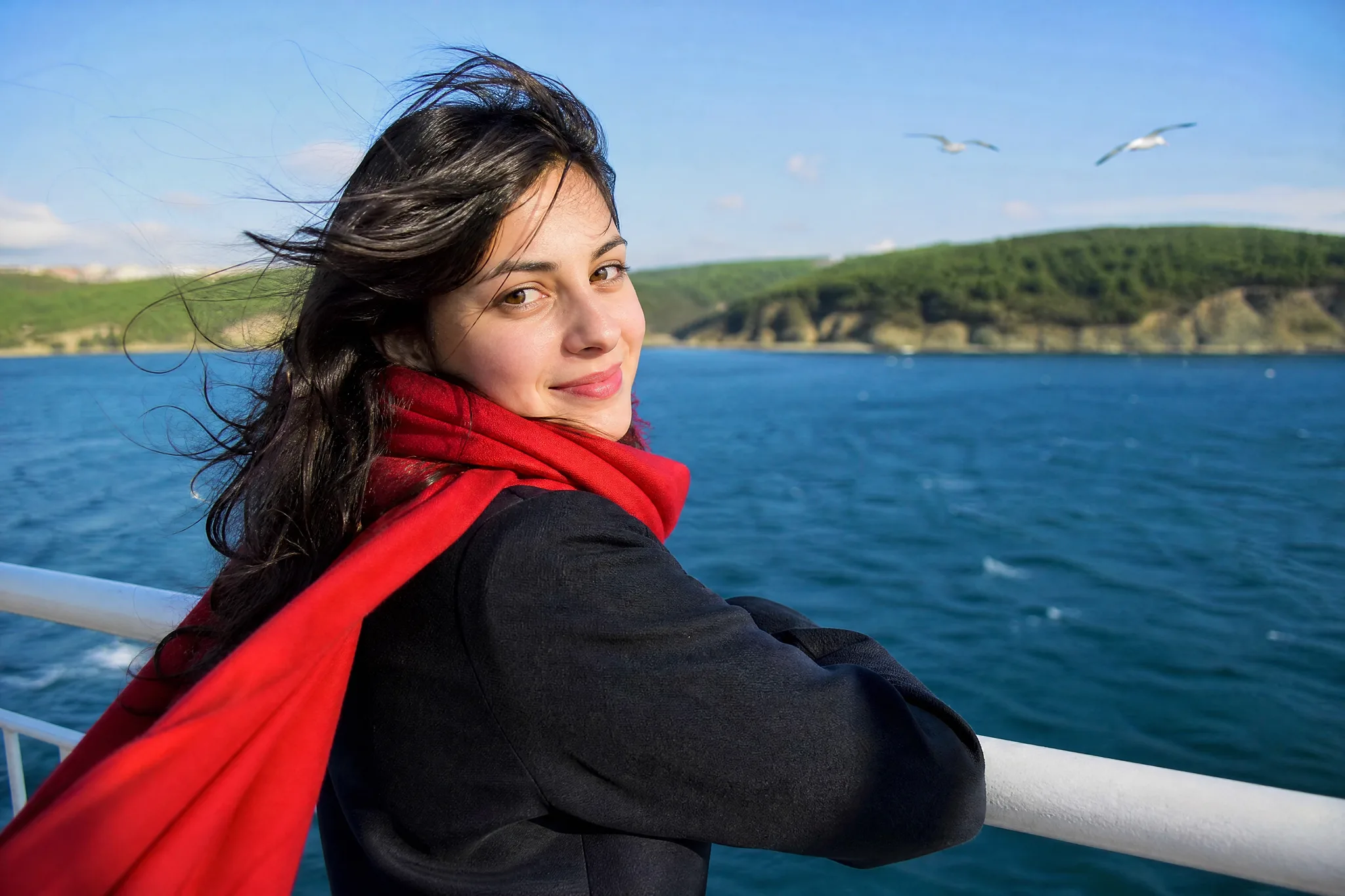 Smiling woman in a red scarf and dark coat looking back over her shoulder at a ferry rail, hair blowing in the sea breeze