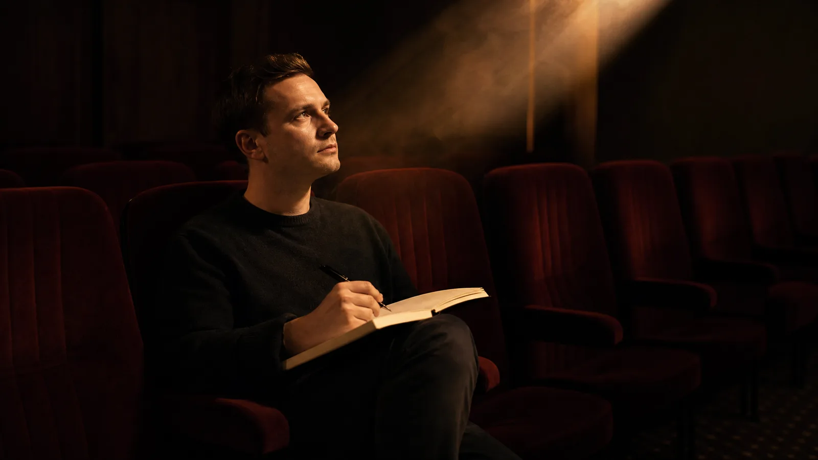 A man in a dark screening room with a blank notebook on his knee, studying a projection beam overhead