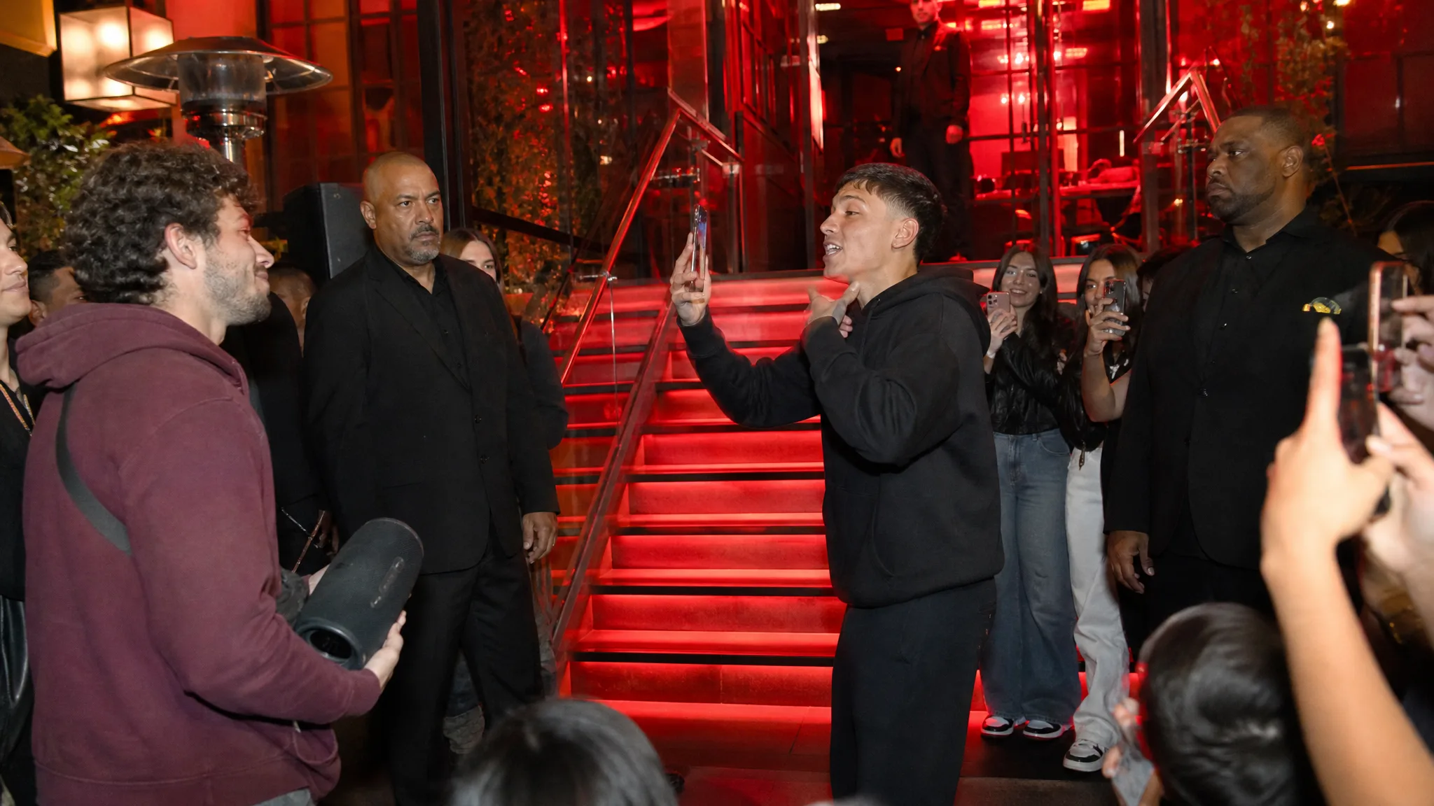 Bulin 47 AI Video Generator: The Venue Scene Wide view of a red-lit entrance with stairs and railings, a performer rapping, a man holding a speaker and fans filming