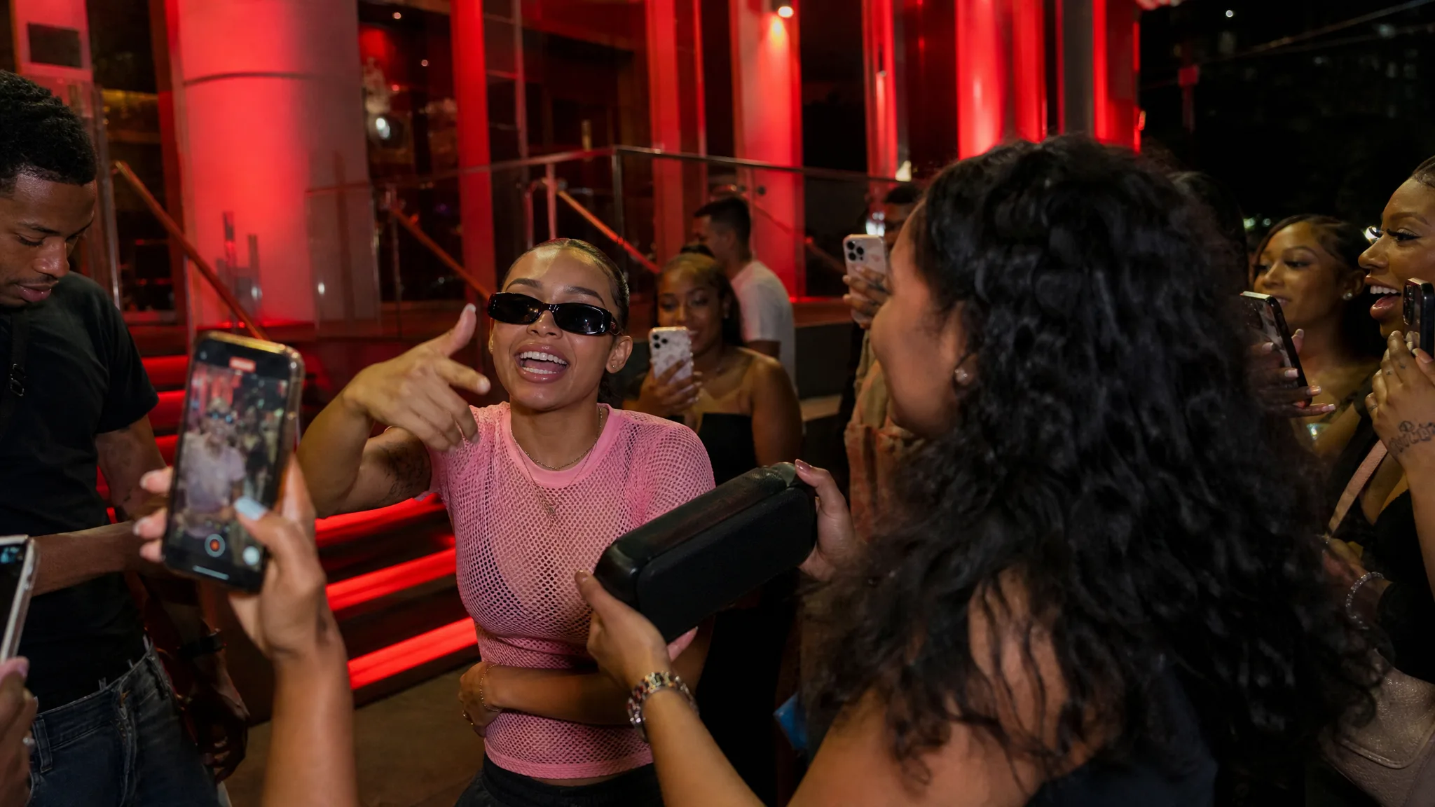 Young woman in sunglasses and a pink mesh shirt freestyling at a red-lit venue entrance as a friend holds a speaker