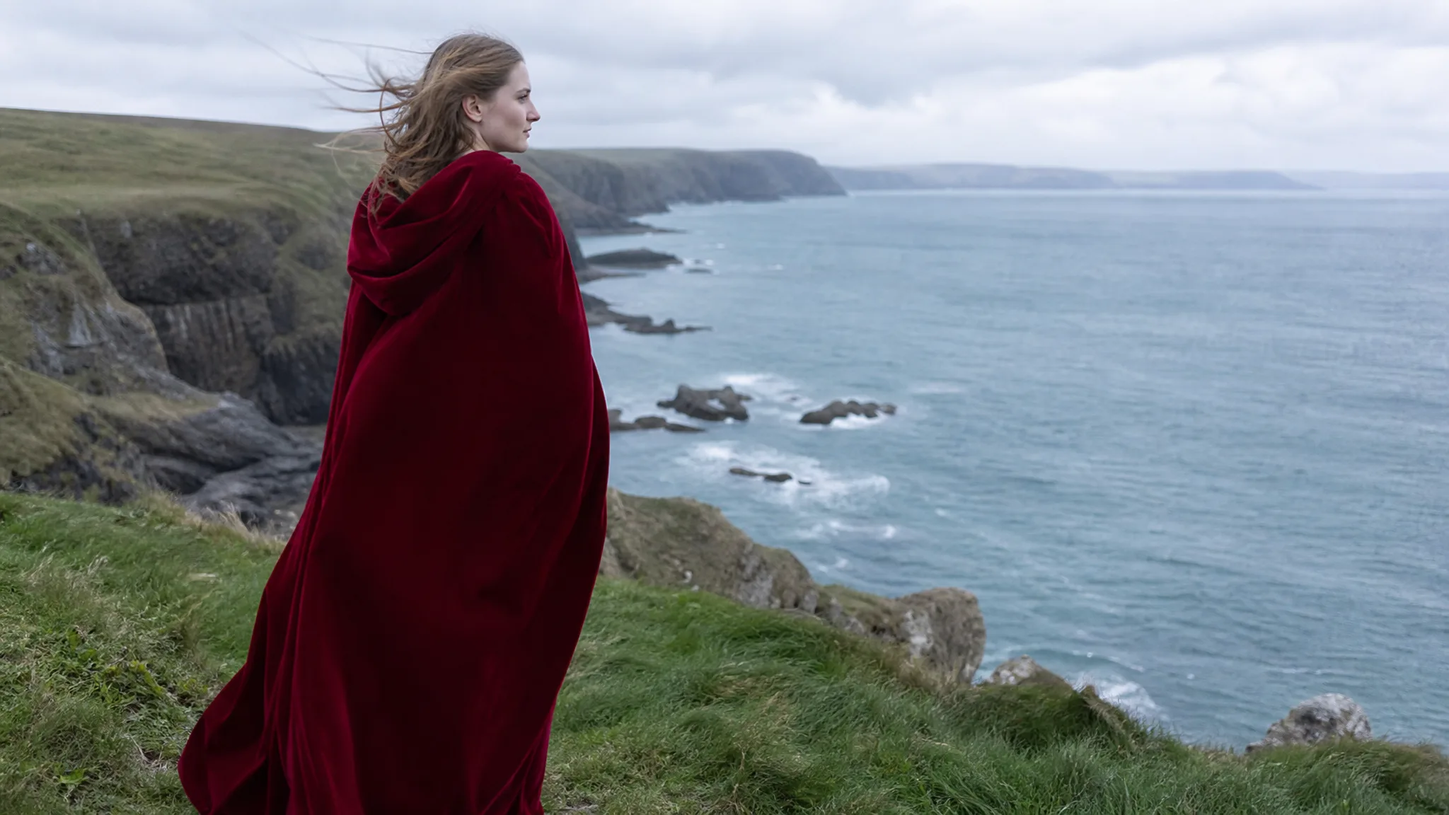 A young white European woman in a long crimson hooded cape stands on a grassy sea cliff under bright overcast daylight