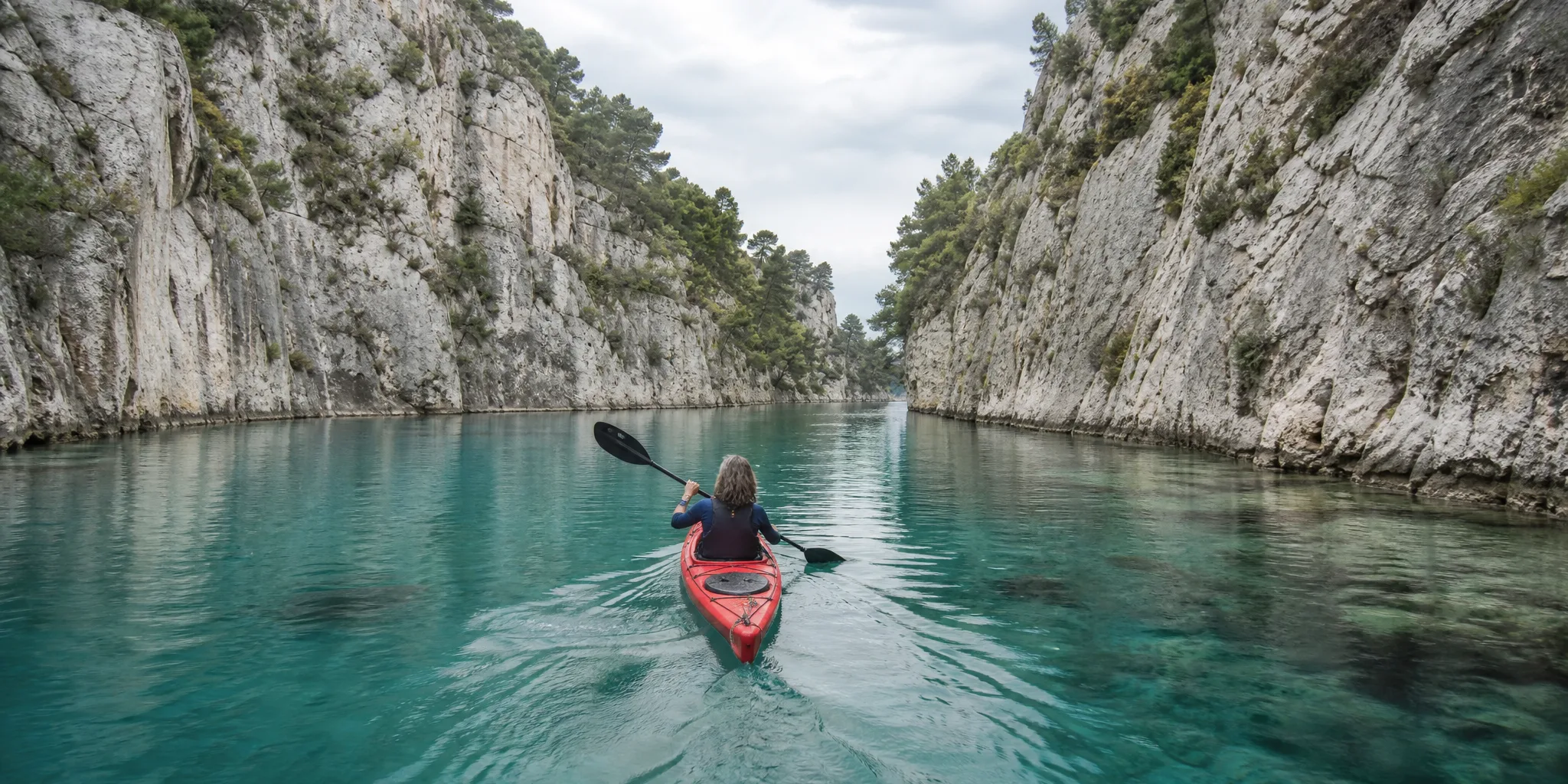 Kayaker in a red kayak paddling through a narrow turquoise gorge between pale limestone cliffs on an overcast day