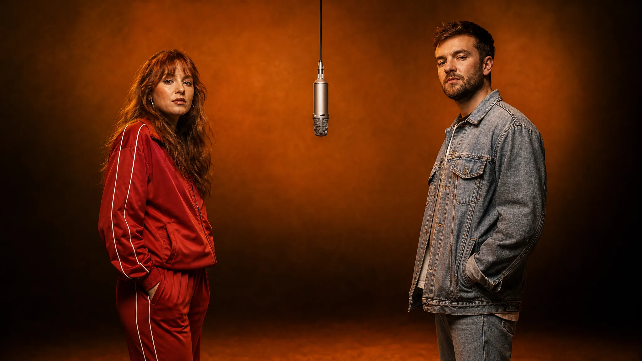 Hotel Lobby AI: One Performer on Each Side Woman in a red tracksuit and man in a denim jacket standing on either side of a hanging microphone in an orange studio