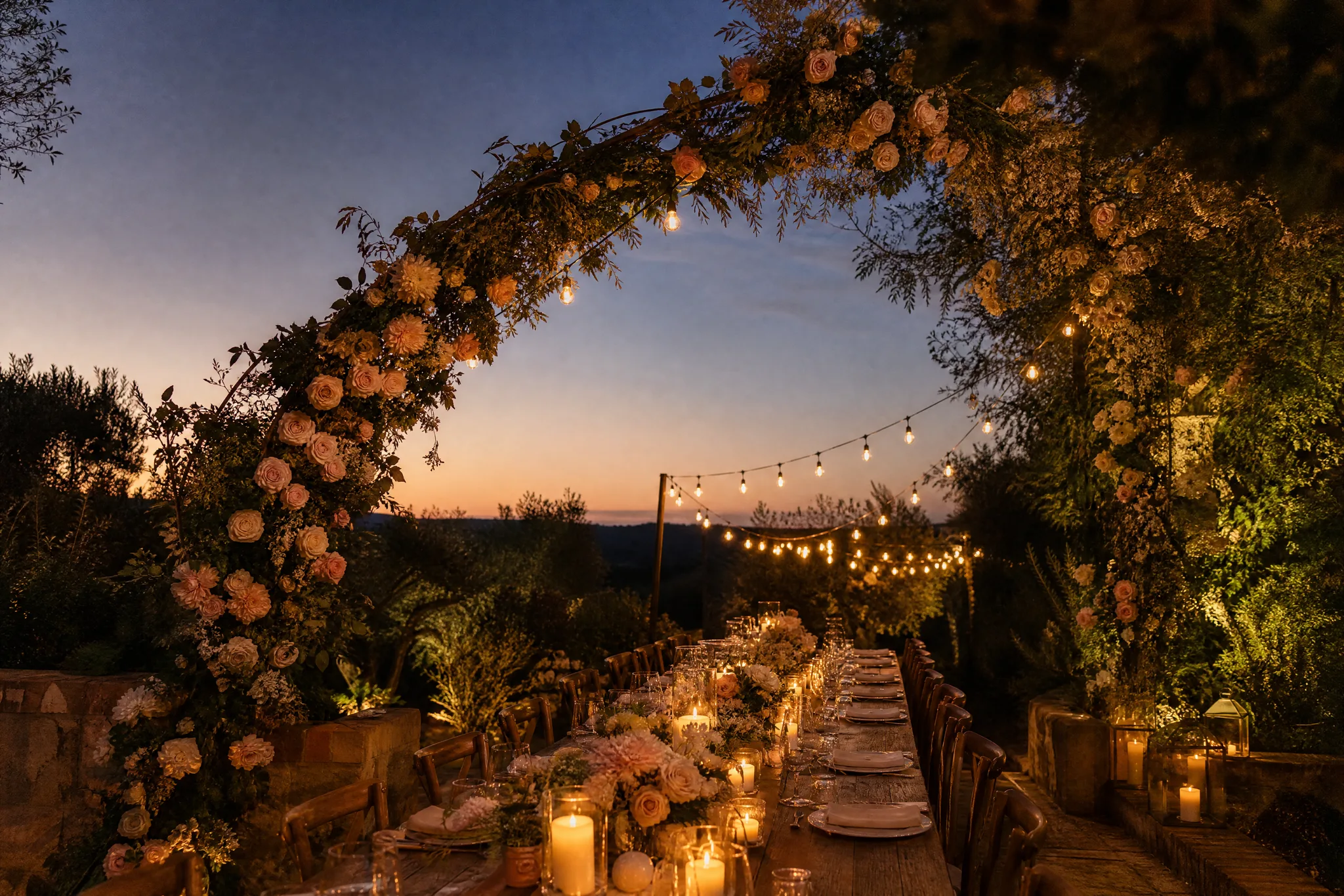Arch of blush roses over a long candlelit garden dinner table at dusk, with open sky above