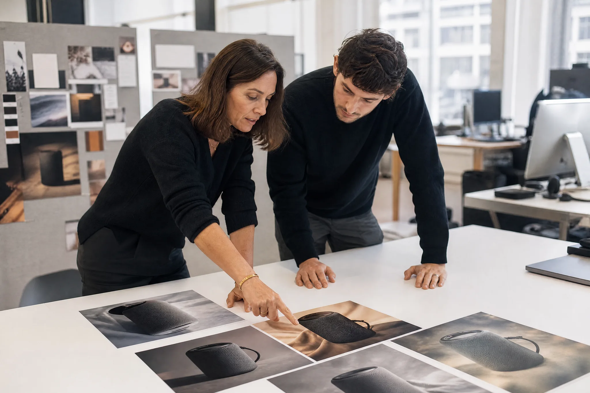 Two colleagues in a bright office comparing printed photos of a charcoal portable speaker laid side by side
