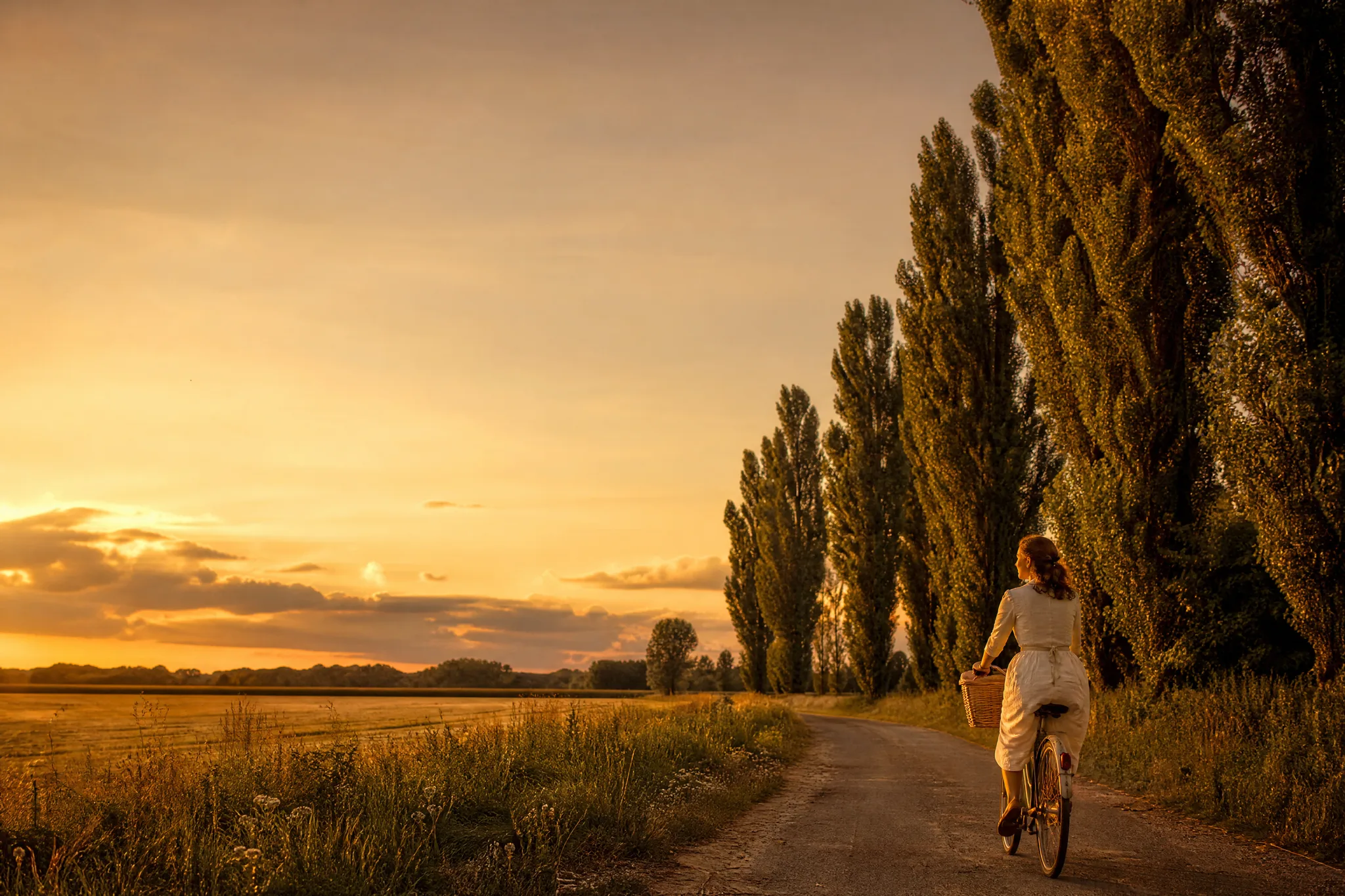 Woman on a vintage bicycle in the lower corner of a country lane scene with open evening sky above
