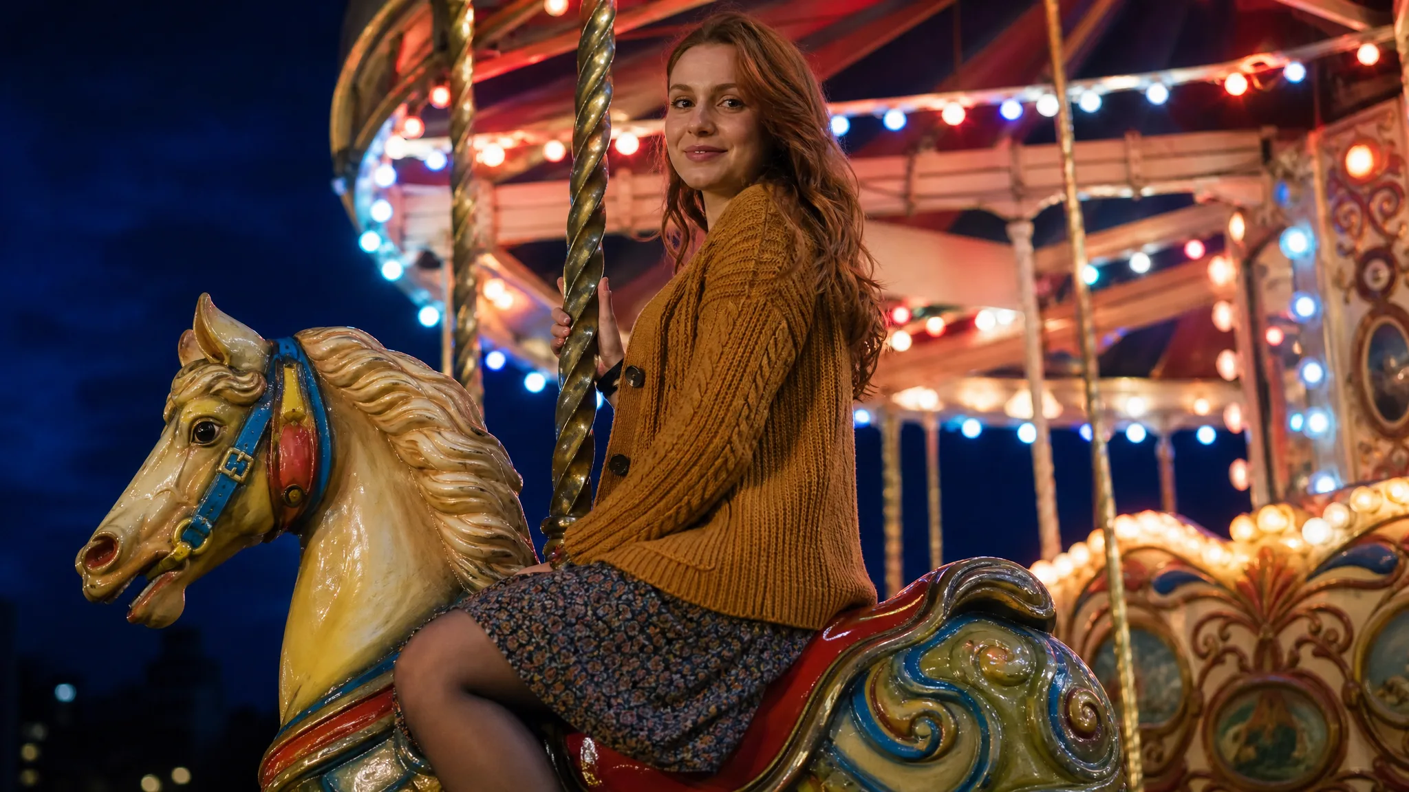 Woman in a mustard cardigan smiling on a painted carousel horse at night, colourful bulbs glowing on the canopy