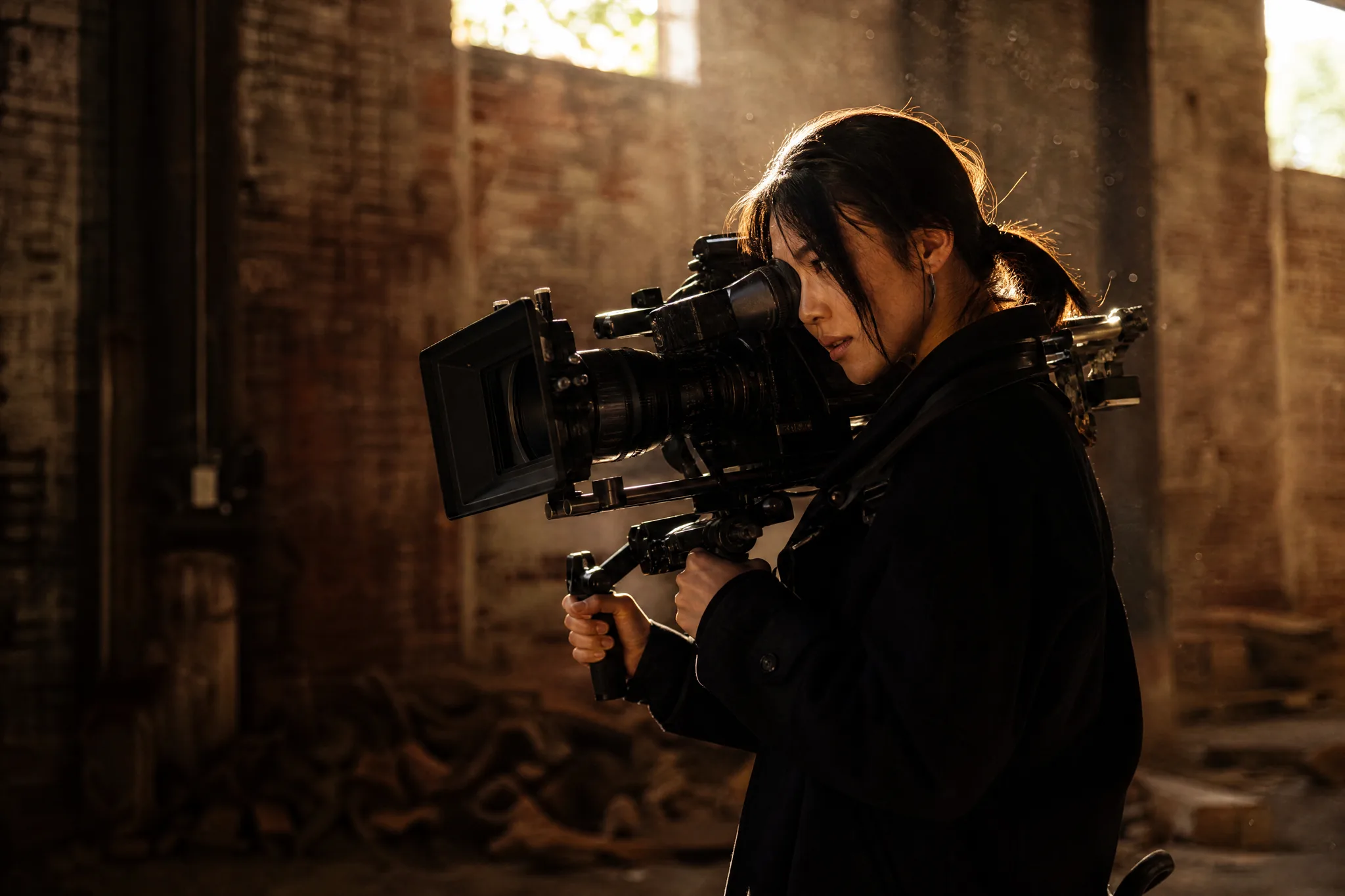A young East Asian woman cinematographer operating a professional cinema camera on a shoulder rig inside a dim brick